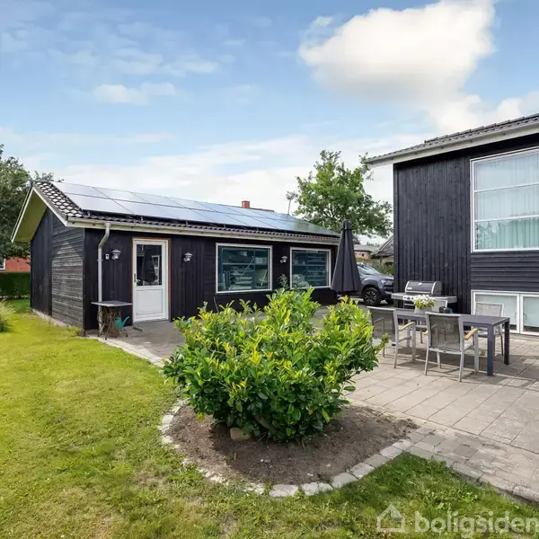 Et sort træhus med solpaneler står ved siden af en flisebelagt terrasse med havemøbler. Huset er omgivet af en velplejet græsplæne og grønne buske under en blå himmel.