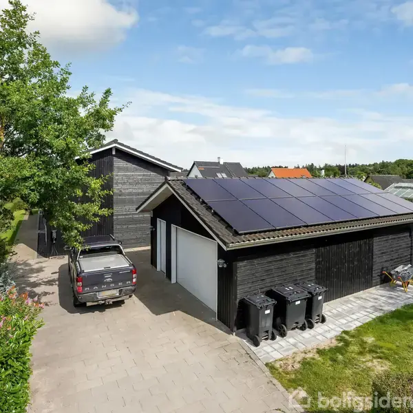 En sortmalet garage med solpaneler på taget står ved siden af en indkørsel, hvor en sort bil er parkeret. Grønne buske og en træstolpe omgiver bygningen.