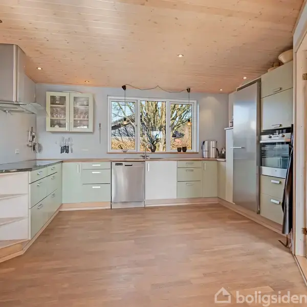 Køkken med lyse skabe, opvaskemaskine og vindue med udsigt til træer. Trægulv og loft med indbyggede lys. Moderne ovn og køleskab ved siden af glasdør til venstre.