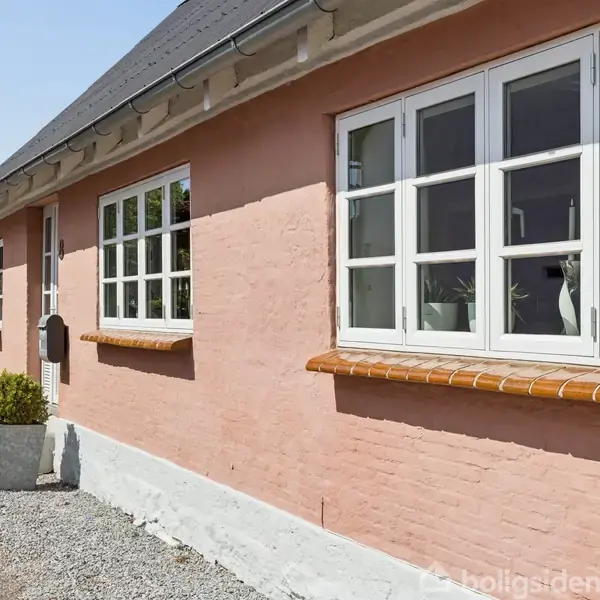 Rosa murstenshus med hvide vinduer står i sollys. Foran huset er der en grusbelægning og grønne planter i hvide krukker.