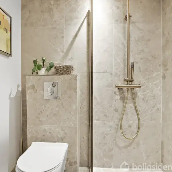 Toilet og bruser i et badeværelse med lys beige marmorklinker. To planter og en kurv står på hylden over toilettet. Toiletpapir er monteret på væggen. En indrammet illustration hænger over toilettet.