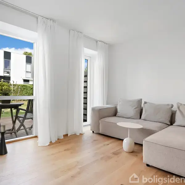 Stuen indeholder en lys beige hjørnesofa med puder, et lille rundt bord, og har udsigt til en udendørs terrasse med træmøbler. Gulv-til-loft-vinduer lukker lys ind.