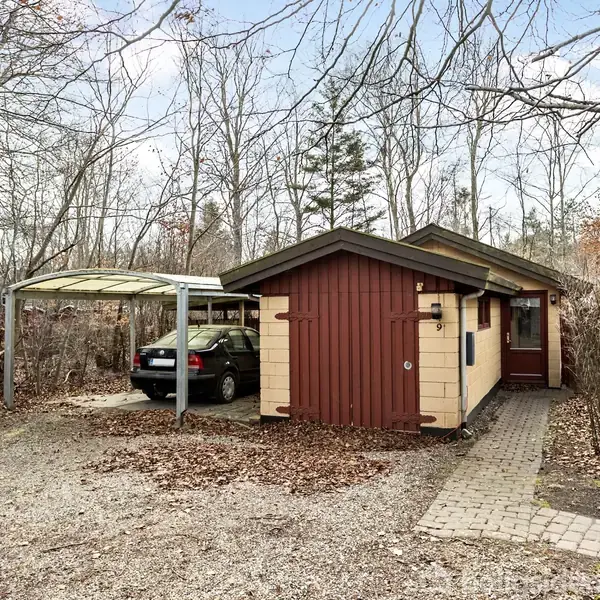 Et lille træhus står stille i en skovklædt have med en grusbelagt indkørsel. En bil er parkeret under en træcarport ved siden af huset.