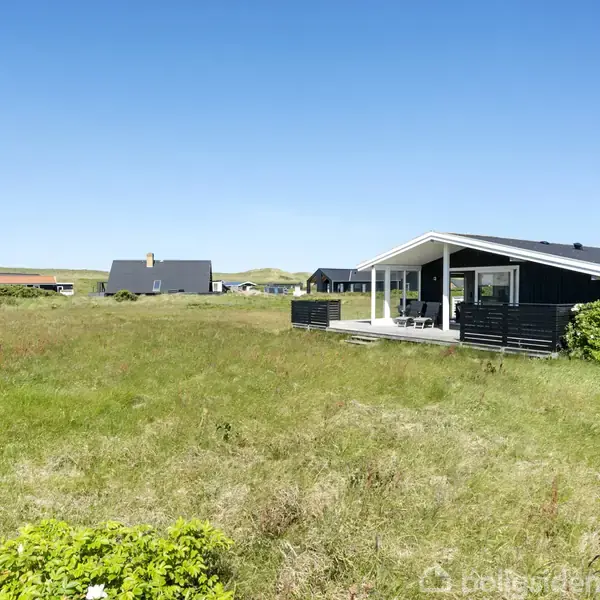 Et sommerhus står stille på en åben grøn eng. Omkring det ses flere huse og sandklitter i baggrunden under en klar blå himmel.