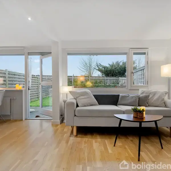 En grå sofa står i en stue ved siden af et rundt sofabord med en plante. Store vinduer og dør åbner ud mod en have. Ingen tekst.