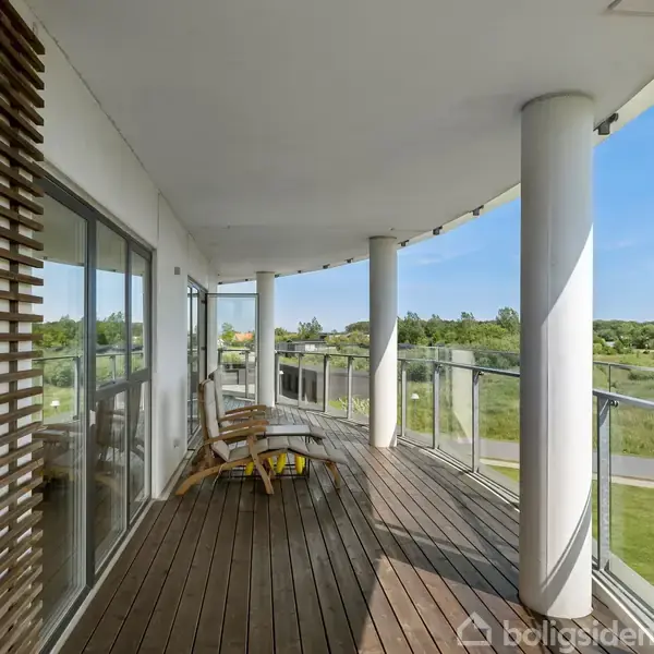 En rummelig altan med trægulv strækker sig langs en hvid bygning. Der står to liggestole, og glasrækværket giver udsigt til et grønt landskab i solskin.