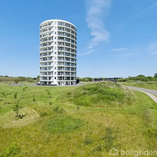 Et rundt, hvidt højhus står stille i en åben, grøn mark med en sti til højre under en klar blå himmel.