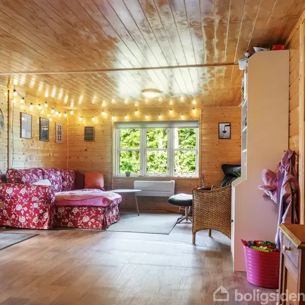 Et træbeklædt rum med sofa i rødblomstret stof placeret ved væggen. Lyskæder hænger fra loftet. Vinduet viser grønne omgivelser udenfor. Et bord og stole omkring rummet.