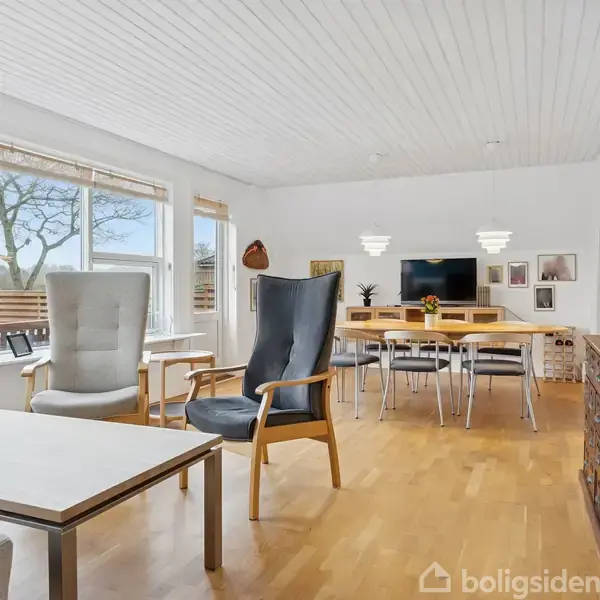 Stue med moderne indretning; to polstrede stole står ved et sofabord. I baggrunden er et spisebord med stole. Rummet har lyse trælofter og store vinduer, der slipper naturligt lys ind.
