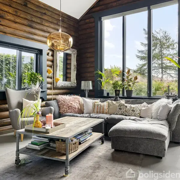 En hyggelig stue med en grå hjørnesofa og et sofabord fyldt med bøger og planter. Store vinduer med udsigt til en have og træpaneler skaber en varm atmosfære.