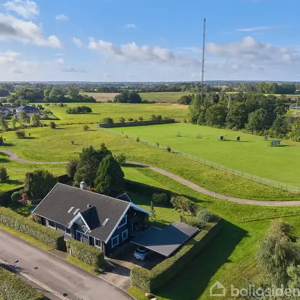 Et hus med mørkt tag og tilknyttet have ligger ved en vej, omgivet af grønne marker og små skovområder under en klar blå himmel.