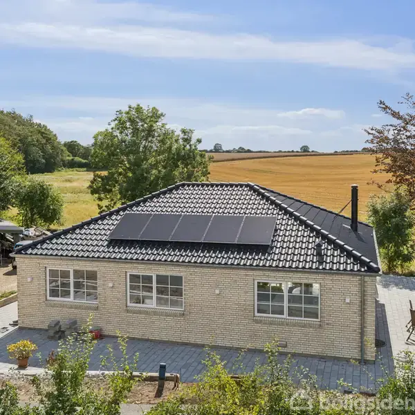 Et hus med solpaneler på taget står i et landligt område omgivet af marker og træer. En terrasse med stole ses ved siden af huset.