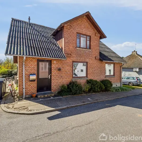 Rødt murstenshus med sort tag, placeret i et villakvarter med en parkeret bil ved siden af. Et skraldespand er til venstre, og sollys oplyser omgivelserne.