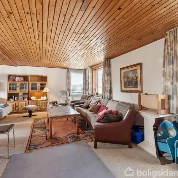 Stue med brunt trægulv og træloft indeholder en stor L-formet sofa, sofabord og reoler. Væggene er dekoreret med malerier, og der er store vinduer med gardiner.