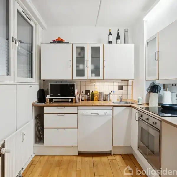 Et køkken med hvide skabe og træbordplade indeholder en ovn, opvaskemaskine og mikrobølgeovn. Overfladen er fyldt med køkkenredskaber og flasker. Vinduer på venstre side giver lys.