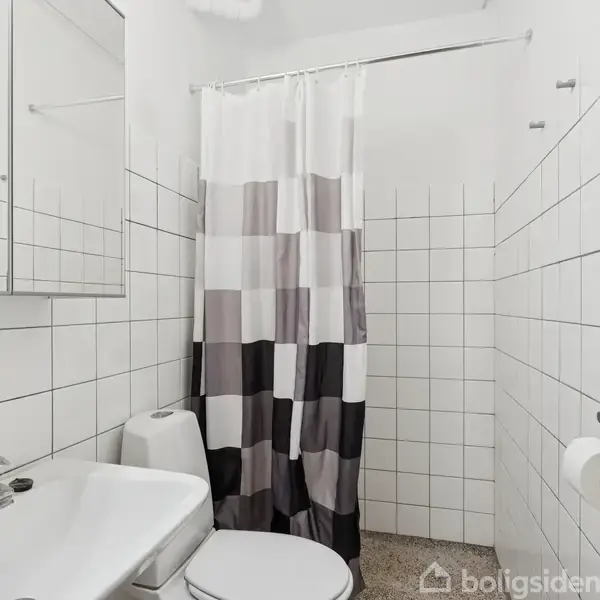 En hvidt flisebelagt badeværelse med vask, toilet og en brusekabine med ternet bruseforhæng. Spejlskab over vasken, toiletrulleholder ved siden af, og gråt gulv.