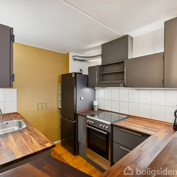 Køkken med mørkegrå skabe og træbordplader. Køleskab, ovn og kogeplade er placeret langs væggen. Vask og vandhane findes til venstre, med en gul væg i baggrunden.