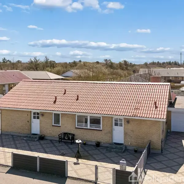 En gul murstensvilla står stille på en flisebelagt gårdsplads. Enkeltstående garager og stakitter omkranser huset i et forstadsområde med flere huse. Klart blå himmel med lette skyer.