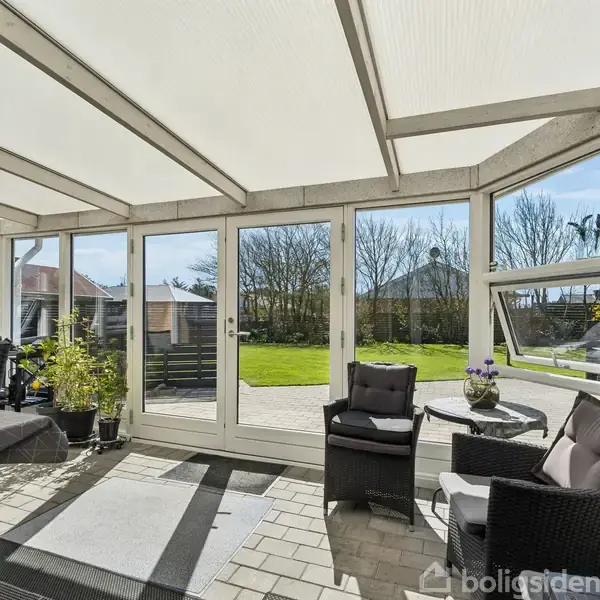 En indglasset terrasse med sorte kurvemøbler og grønne planter. Døren åbner ud mod en solrig have med græsplæne og træer. Klar himmel ses gennem glaspanelerne.
