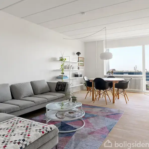 Grå sofa med tæppe står i stue, sofabord med pyntegenstande foran. Til højre spisebord med sorte stole. Store vinduer giver udsigt og lysindfald. Hylder og dekoration hænger på væggen.