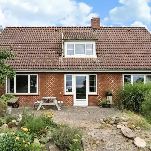 Rødt murstenshus med skråtag og et frontvendt kvistvindue. Foran huset er der en flisebelagt terrasse med stenbed og planter. Bag ved er der nogle træer i den grønne have.