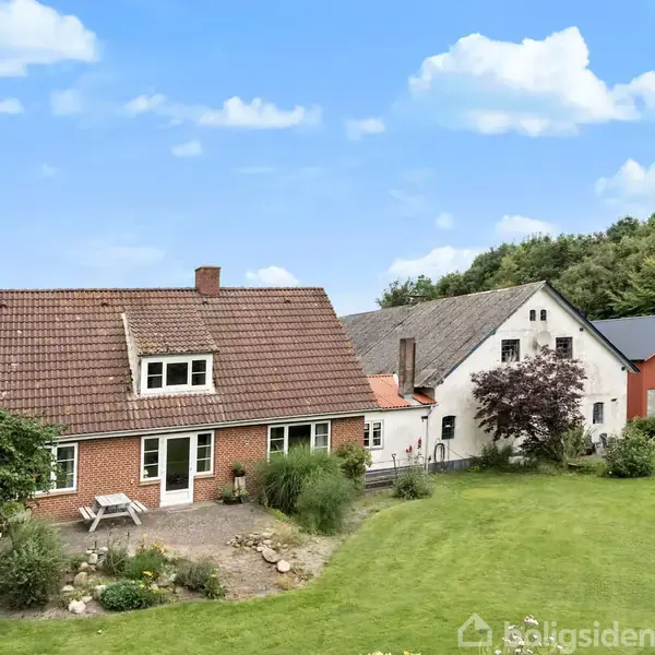 Et rødt murstenshus med skråt tag står i en grøn have. Bagved ses en hvid lade, og der er træer i baggrunden under en blå og skyfyldt himmel.