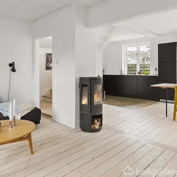 En moderne stue har en tændt brændeovn, placeret på lyse trægulve. En sort sofa står ved et rundt træbord med bøger og stearinlys. Køkkenet med sorte skabe ses i baggrunden.