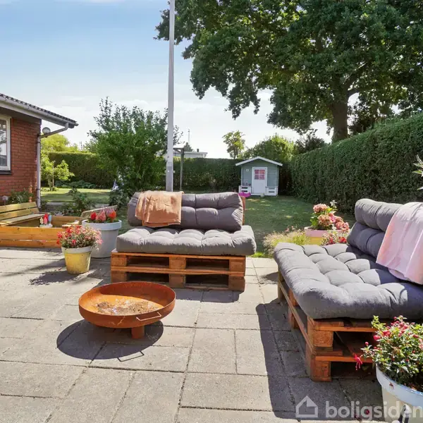 To udendørs pallesofaer med grå hynder står på en terrasse foran et rødt murstenshus, omgivet af blomsterkrukker og et bålsted, med en have i baggrunden.