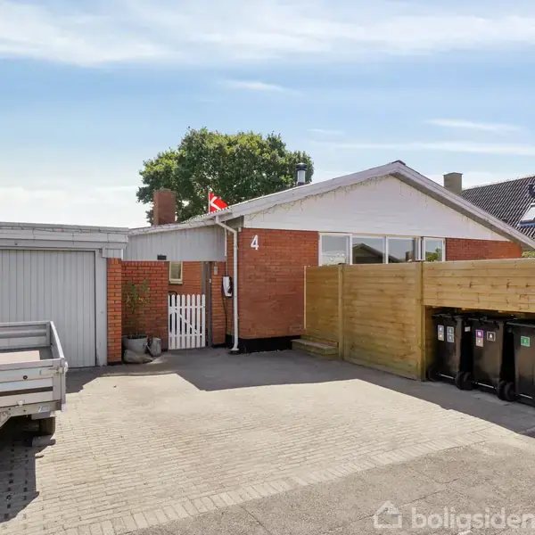 En rød murstensbygning med et fladt tag og en flagstang. Nummeret "4" er synligt ved indgangen. En trailer står parkeret på en brolagt indkørsel ved siden af en lukket garage.