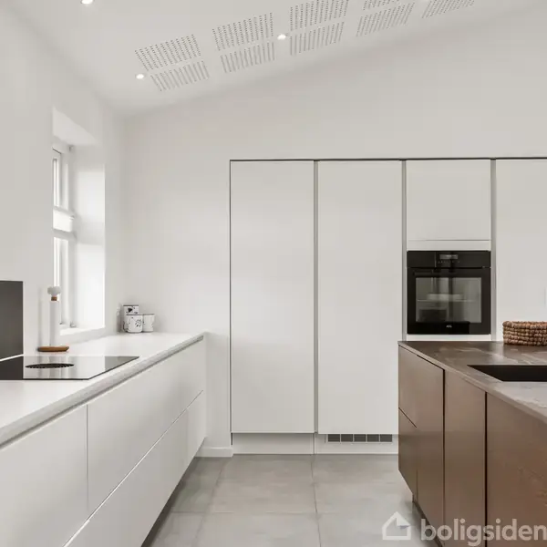 Køkken med integrerede hvide skabe og sort vask. Indbygget ovn og moderne kogeplade. Rummelig og lys atmosfære med minimalistisk design og store, lyse fliser på gulvet.