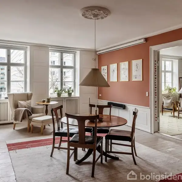 Et spisebord med fire stole i en stue med røde vægge. Et loftslys hænger over bordet. Der er to vinduer og billeder på væggen. Adjacent er en anden stue med reoler.