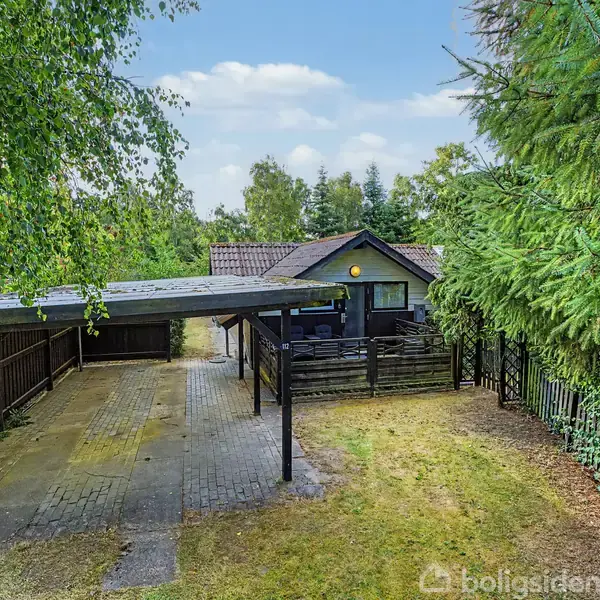 Et lille træhus står i en have omkranset af grønne træer. Foran huset er der en carport med belægningssten. Huset har en terrasse med en lampe over døren.
