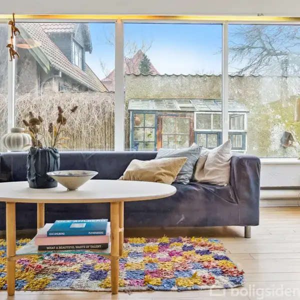 En mørkeblå sofa står i en stue foran store vinduer med udsigt til en have. En træbord med bøger og en skål står foran sofaen, på et farverigt tæppe.