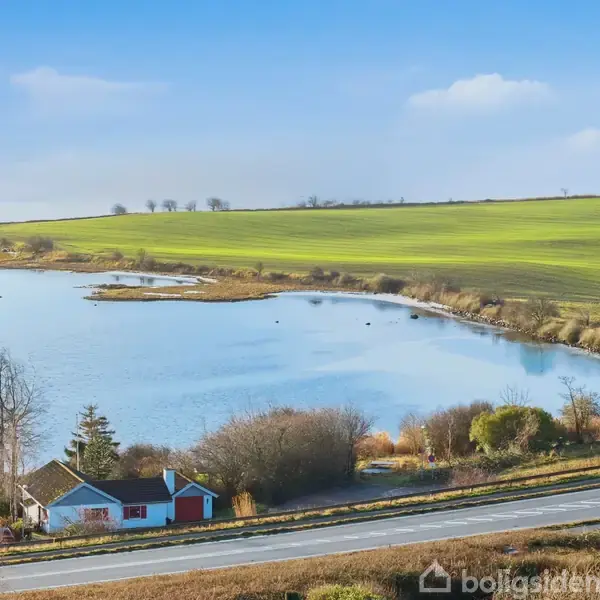 Hus ved en vej med udsigt over en sø, omgivet af marker og buske. Blå himmel i baggrunden afsluttes ved horisonten af lave træer.