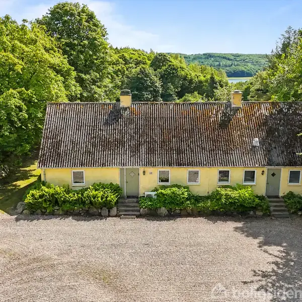 Gult hus med mosdækket tag står i skovområde, omgivet af træer. Huset har flere indgangsdøre og vinduer med småstensbelagt forplads. Baggrunden viser udsigt til bakker og skov.