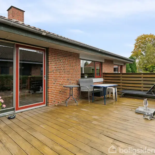 Terrassen har træplanker og er møbleret med et bord og stole. Den støder op til et murstenshus med store vinduer. I baggrunden er der træer og en flagstang.