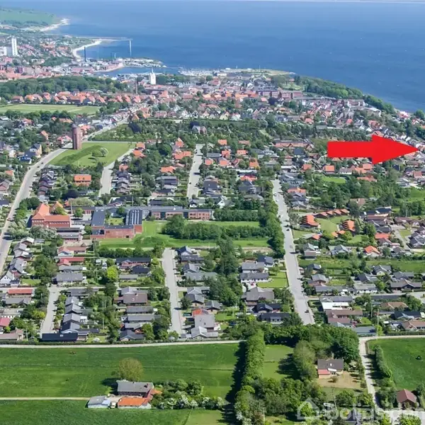 Røde pil peger mod hus i forstad. Mange huse omgivet af træer i et område nær kysten. Byen strækker sig mod havnen med åbne grønne områder i forgrunden.