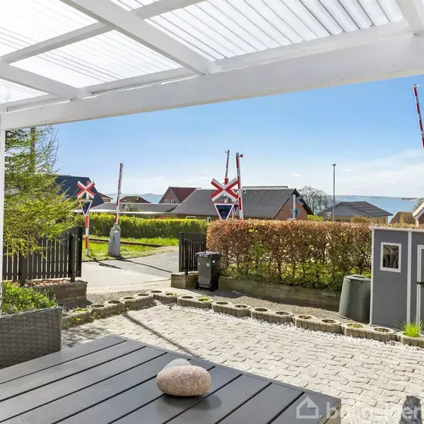 En overdækket terrasse med grå sofa og bord står i forgrunden. I baggrunden ses et roligt nabolag med hække, træer, et hegn, og en udsigt over vandet.