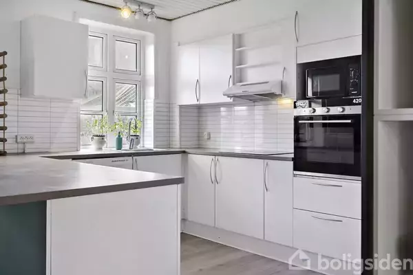 Et moderne køkken med hvide skabe og apparater, herunder en integreret ovn og mikrobølgeovn. Der er planter på vindueskarmen, og rummets vægge har hvide fliser.