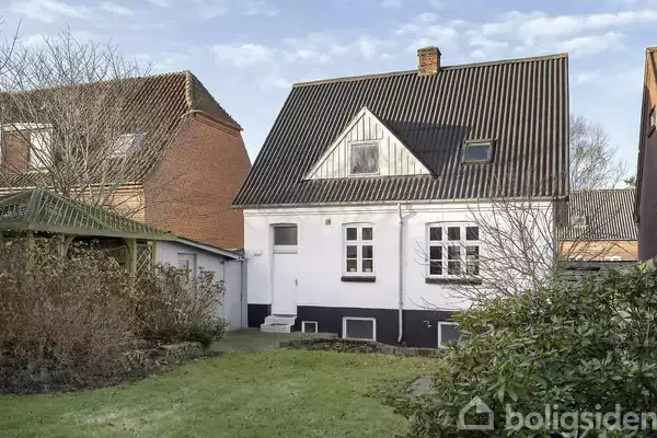 Et hvidt hus med sort tag har en hoveddør og vinduer mod baghaven. Omkranset af buske og træer står det mellem to andre huse.