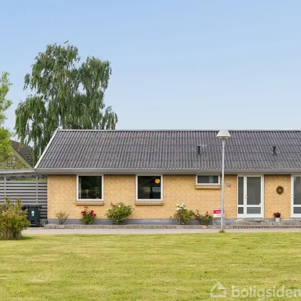 Et gult murstenshus står foran en velplejet græsplæne. Huset har et gråt tag, vinduer og en havedør. Der er blomster foran huset, og en lygtepæl står i nærheden.