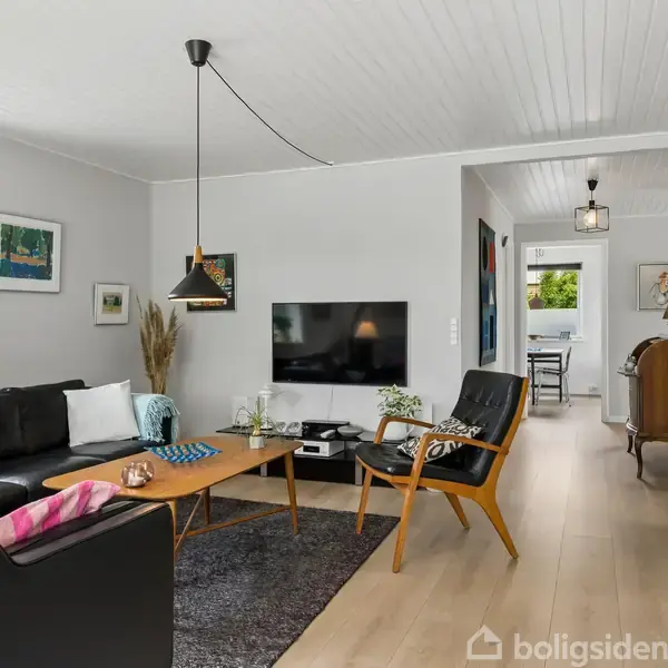 Stue med sort lædersofa og træstole omkring et lavt sofabord. Fjernsyn hænger på væggen, bøger står på hylder. Store vinduer lader lys komme ind.