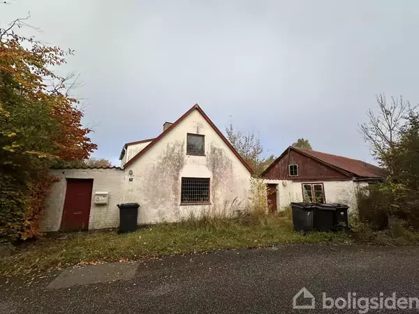 En hvid bygning med rødt tag står i et overgroet område. To affaldscontainere er placeret foran huset. Omgivelserne er fyldt med træer og lavt græs.