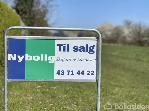 Skilt står i en have og annoncerer "Nybolig Til salg Skiffard & Simonsen 43 71 44 22". Det er omgivet af grøn græsplæne og træer i baggrunden under en blå himmel.