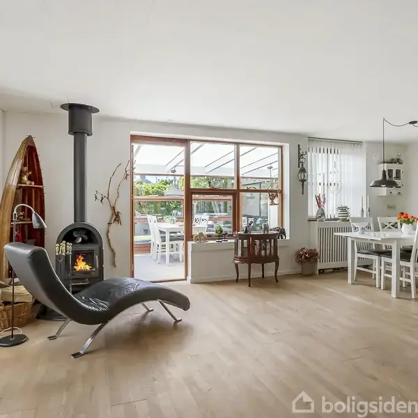 Brændeovn står i stue med trægulv og moderne lænestol. Store vinduer giver udsigt til en overdækket terrasse. Et spisebord med seks stole ses til højre for rummet.