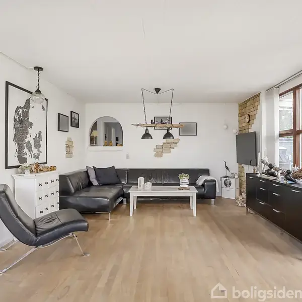 Sort læderhjørnesofa står i en stue med lyse trægulve, flankeret af et sofabord og et skab. Store vinduer til højre giver udsigt til udendørsgrønt. En sort lænestol står til venstre.