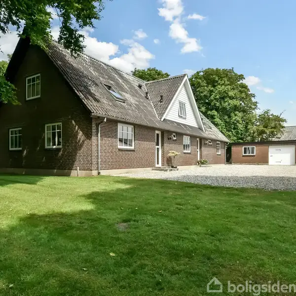En rødstensvilla med spidst tag står foran en græsplæne. Bygningen har flere vinduer og en tilhørende garage med to porte. Himlen er klar og solrig.