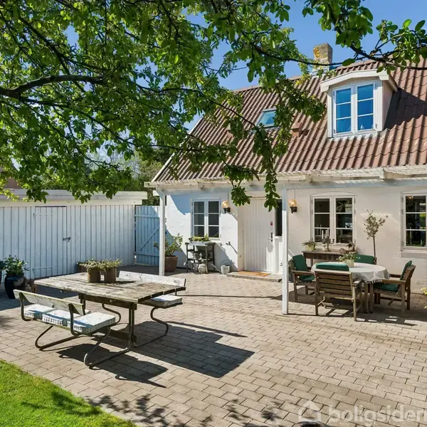 Et hyggeligt gårdhavemiljø med en hvidmalet murstensbygning. En terrasse med borde og stole under et træ, omgivet af grønne planter, skaber en afslappet atmosfære.