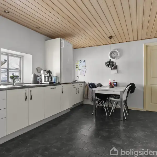 Et moderne køkken med hvide skabe og sorte bordplader. Ved et rundt bord står fire stole. Et vindue og en dør ses i baggrunden under et træloft.