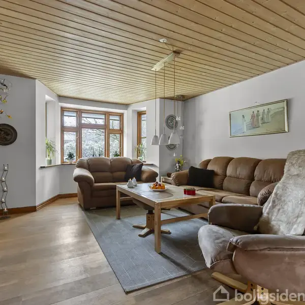 En stue med brune lædersofaer rundt om et træbord, dekoreret med pynt og planter på vindueskarmen. Rummet har lyse vægge, træloft og forskellige billeder ophængt.